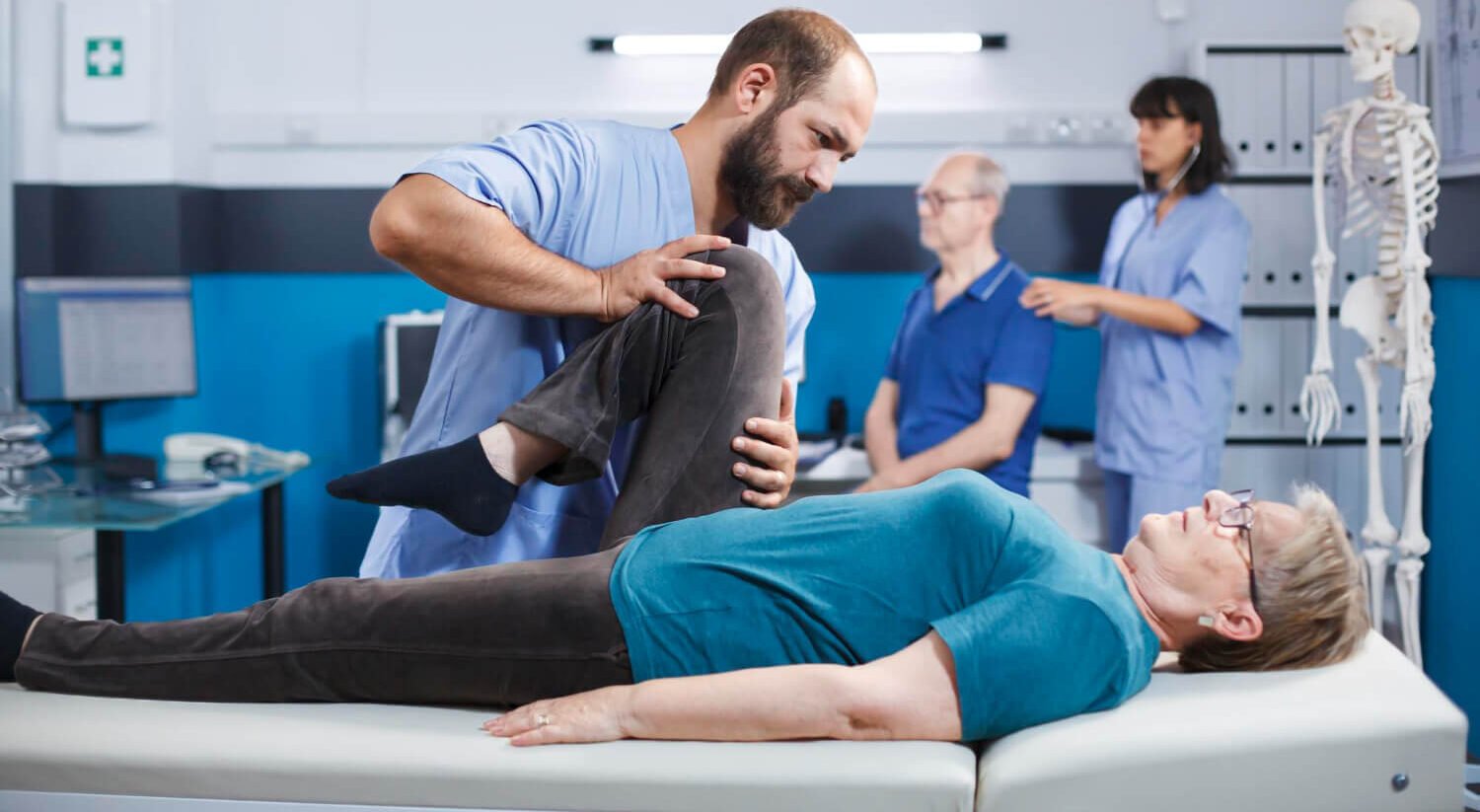 A physical therapist helps an older woman with a leg stretch on an exam table. In the background, two other people consult while a skeleton model stands nearby in a clinical setting.