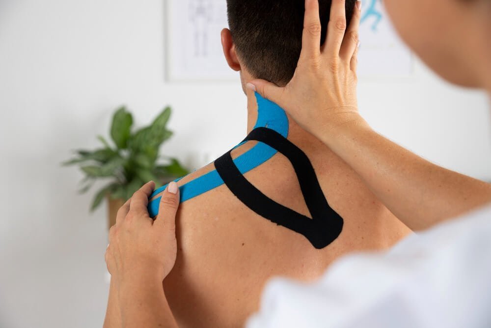 A person applies blue and black kinesiology tape to the back and shoulder of another person, who is sitting shirtless, during a physical therapy session. A green plant is visible in the background.