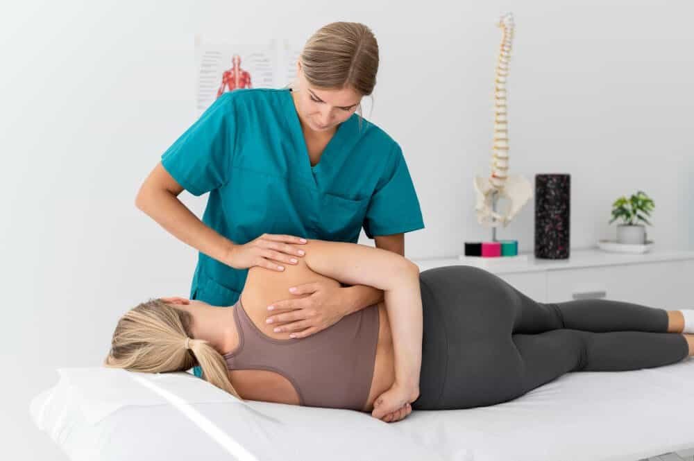 A physical therapist in teal scrubs adjusts the back of a woman lying on her side on an exam table in a bright clinic, with a spine model and medical chart visible in the background.