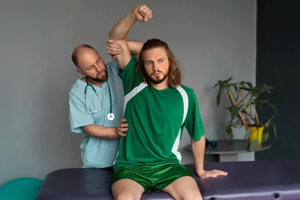A healthcare professional in scrubs assists a man in a green sports outfit with a shoulder stretch on an exam table in a clinic room with plants in the background.